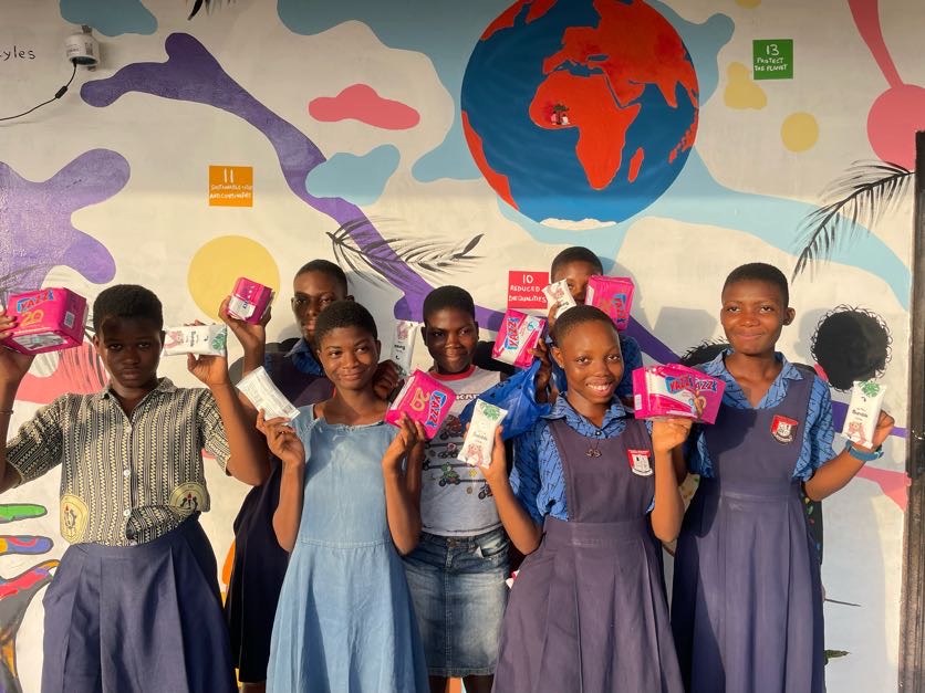 Smiling schoolgirls in uniform hold up donated care packs in front of a colourful world mural.